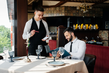 A young businessman in a fine restaurant examines the menu and makes an order to a young waiter in a stylish apron. Customer service. Table service in the restaurant.