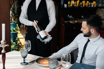 A young sommelier in a stylish uniform demonstrates and offers the client fine wine in the restaurant. Customer service. Table setting in a fine restaurant. - obrazy, fototapety, plakaty