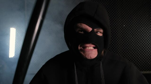An angry man in a balaclava and hood is standing with a bat and shaking his fist. The concept of robbery and protests
