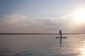 Silhouette of a beautiful woman swimming on big lake staying Paddle SUP Board at sunset background.