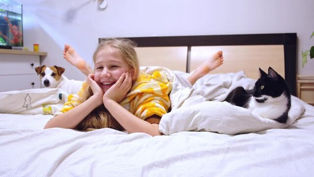 Laughing blonde teen girl lying on bed with cat and dog watching TV at home. A happy child is relaxing at home and watching movie or cartoon, resting his head in his hands. Cozy home concept.