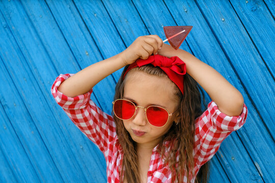 A girl with a red bow on her head and sunglasses holds a triangular shaped lollipop in hands. The child looks into the camera. Blue old painted Background from planks