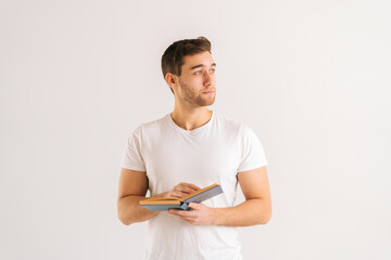 Studio portrait of focused young man holding in hands paper book on white isolated background in studio, looking away. Front view of serious male student studying reading educational materials.