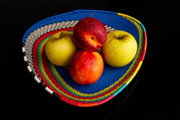 A colourful fruit basket with apples and peaches on top of a reflective black surface. 