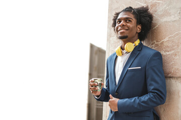 smiling young black afro ethnicity businessman with coffee and headphones leaning on wall, copy space