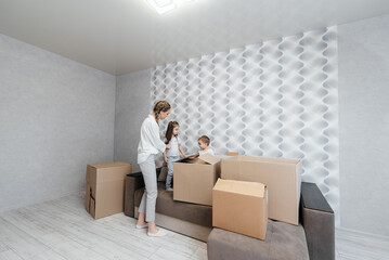 A young mother with children unpacks a box of things after moving to a new apartment. Purchase of real estate. Housewarming, delivery and transportation of goods.