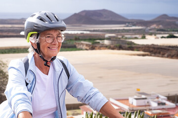 Attractive smiling adult mature senior cyclist woman riding in a country road, cyclist female wearing sport helmet looking ahead. Sport activity healthy lifestyle for retired people.Horizon over water