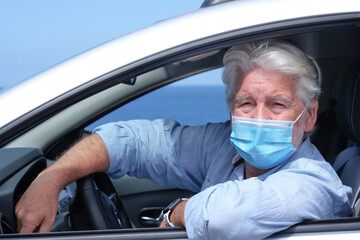 Senior adult man wearing surgical mask due to coronavirus covid 19 omicron variant and delta variant sitting inside the car. Elderly white haired grandparent looking at camera, horizon over sea