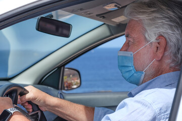 Senior adult man wearing surgical mask due to coronavirus covid 19 omicron variant and delta variant sitting inside the car driving. Elderly white haired grandparent looking ahead, horizon over sea