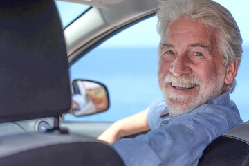 Beautiful senior white-haired man inside his car smiles looking at camera. Horizon over water
