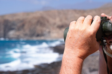 Close up of senior man hands holding binoculars looking at sea, mountain. Senior retired enjoying freedom and holiday