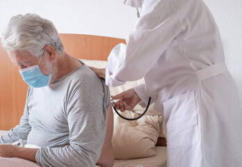 Coronavirus covid 19. Doctor listening to an elderly man's breathing in bed during a home visit....