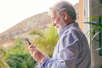 Smiling attractive adult mature senior man standing at the window holding a smartphone enjoying tech and social. Elderly white haired in relaxed moment using cellphone applications. Copy space