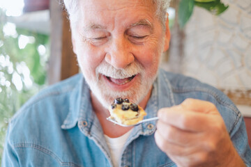 Adult senior bearded caucasian man sitting in cafe eating a piece of fruitcake with cream and...