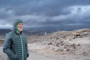 Smiling active adult mature senior bearded man hiking outdoors on a windy cloudy day covering his...