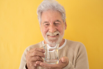 Defocused attractive bearded adult senior man holding a glass of fresh water standing on yellow background. Bearded elderly smiling ecologist man