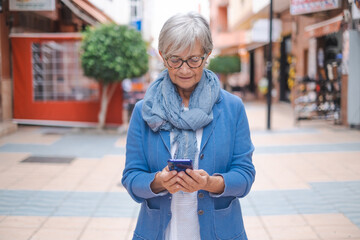 Attractive relaxed mature adult Caucasian woman with gray hair walking in city street holding cellphone in hand. Beautiful senior woman blue dressed enjoys free time and smiles looking at smart phone © luciano