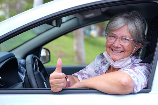 Happy adult attractive caucasian senior woman taking off the surgical mask due to coronavirus. Elderly smiling female sitting inside the parked car looking at camera with thumb up