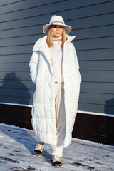 Young woman posing in total white fashion sport chic look with long down jacket, wool costume and...