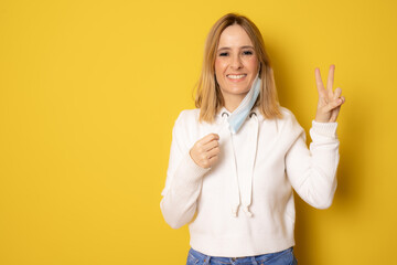 Beautiful woman taking her surgeon mask off isolated over yellow studio background.