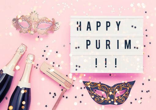 Purim concept. Happy Purim in light box, champagne, carnival mask and gragger on pink background