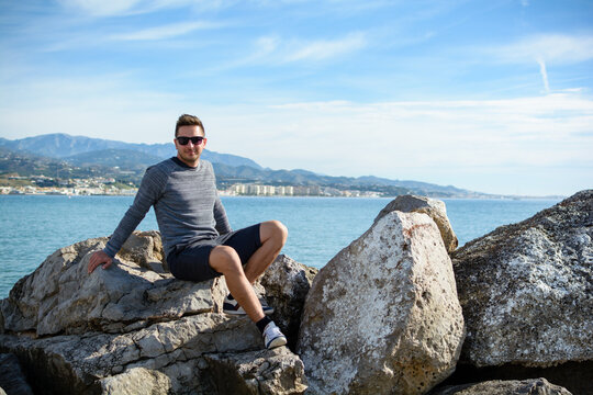 Junger Mann sitzt glücklich auf einem Stein im Urlaub an der Costa del sol in Spanien am Mittelmeer