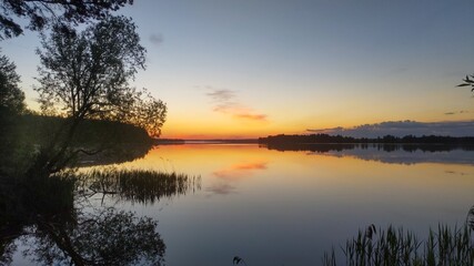 Fototapeta na wymiar The sun rises over the lake on a spring morning. The shores of the lake are covered with forest, reeds grow in the water Tree branches hang over the water The sky and clouds are reflected in the water
