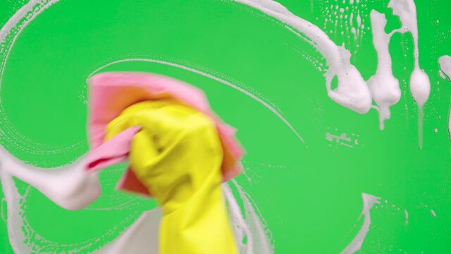 Woman's hand in rubber yellow glove washes window on green screen chroma key background. Housekeeper wipes soap suds from the glass. Cleaning windows with cleaning disinfectant. Close up.