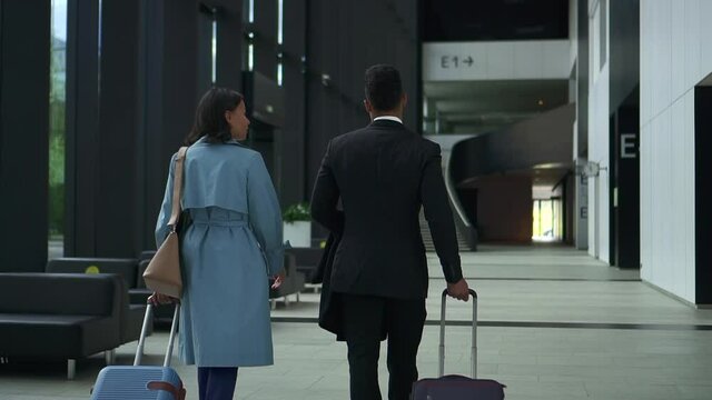 Back view of businesspeople talking and walking with suitcases in hand at office or terminal spbd. American African businessman, businesswoman talk and look with smiles, move luggage and walk in