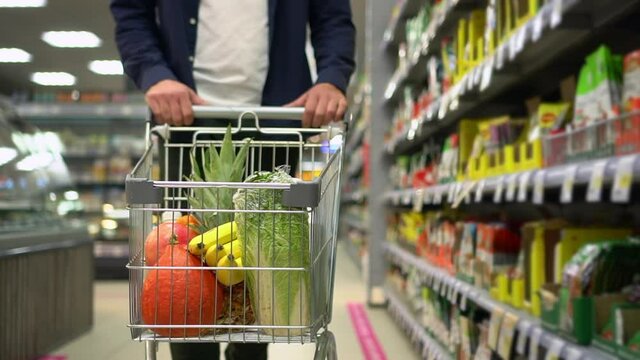 Man consumer choosing organic products and walking with cart in hands at supermarket or market spbd. Closeup view of young male shopper moves trolley with fresh fruits, vegetables and walks along