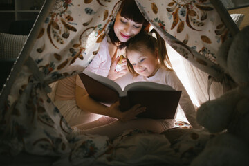 Caring caucasian mother sitting with her daughter inside toy wigwam and using flashlight while reading fairytale. Dark atmosphere at cozy home. © sofiko14
