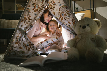 Beautiful woman and pretty child smiling on camera while sitting in wigwam with flashlight and book. Happy mother with daughter spending evening time together at home. © sofiko14