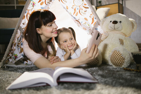 Playful caucasian woman and female child using modern smartphone for taking selfie inside toy wigwam. Mother and daughter playing together during evening time at home.