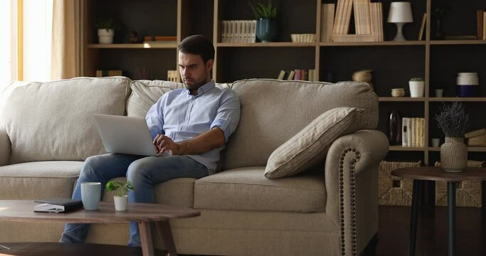 Serious young man typing on laptop keyboard sit on sofa at home, looks pensive, concentrated browsing or chatting on internet. Millennial guy working studying on wireless computer, surfing web concept