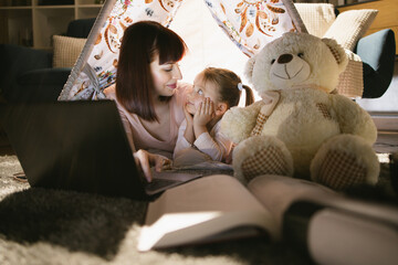 Mother and daughter using wireless laptop while playing together in toy wigwam during evening time. Happy caucasian family with technology at home. © sofiko14