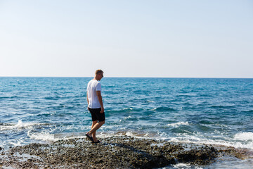 Young attractive man in sunglasses in a white t-shirt and shorts stands on the shore of the mediterranean sea