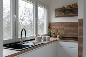 Kitchen sink with faucet near the window. White modern furniture and wooden tiles on the wall in...