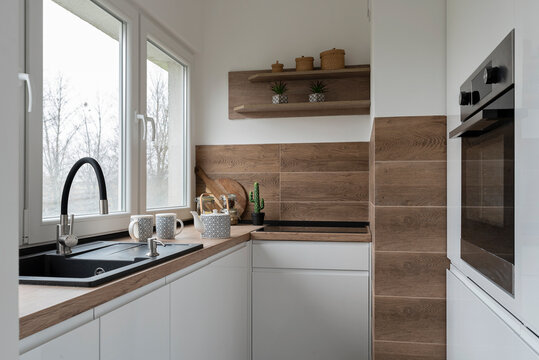 Modern kitchen interior with kitchen sink near the window and wooden tiles on the wall in stylish design. White furniture and oven  in small kitchen.