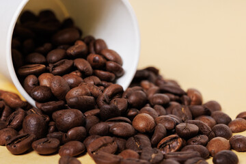 Arabica coffee beans scattered from a white paper cup on a yellow background.