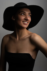 Dark studio fashion portrait of elegant young and sexy woman in black wide hat and black classic evening dress. Stylish elegant woman with beautiful smile and look aside of camera in grey background