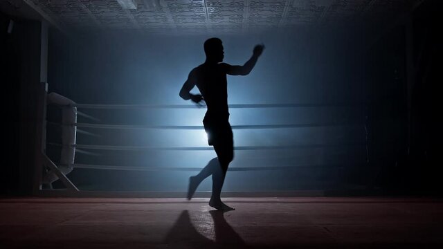 Kickboxer training in low light gym. Muay thai fighter practicing shadow boxing. Male boxer training on dark smoky background with light. Unrecognizable man. Full length in 4K, UHD