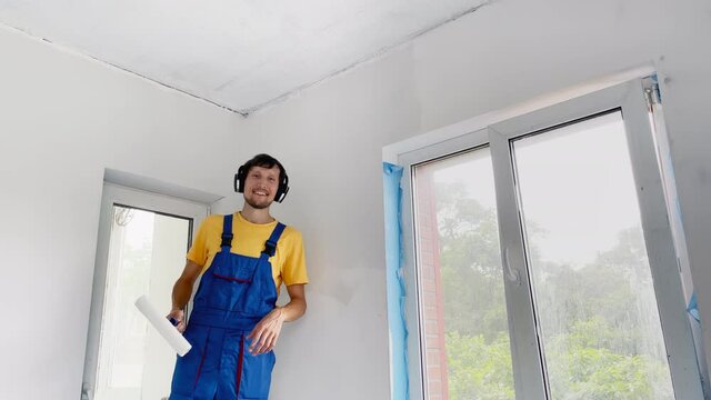 A man professional construction worker paint the wall using a roller painter. He listens to some music in his headphones Slowmotion shot