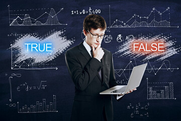Attractive thoughtful young businessman holding laptop with true and false mathematical theories on chalkboard. Science and education concept.