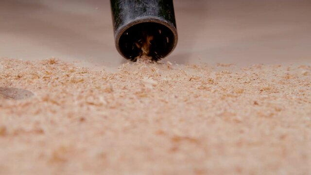 SLOW MOTION, MACRO, DOF: Black plastic vacuum cleans up wood particles lying on the ground. Black vacuum cleaner hose sucks up wood chippings and sawdust scattered across the carpenter's workbench.