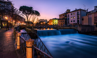 Fototapeta na wymiar Treviso, the San Martino Bridge at sunset. On the right a little waterfall on the Sile river, on the left a walk illuminated by street lamps