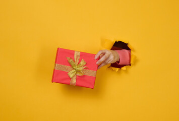 female hand holds a box with a gift on a yellow background, part of the body sticks out of a torn...