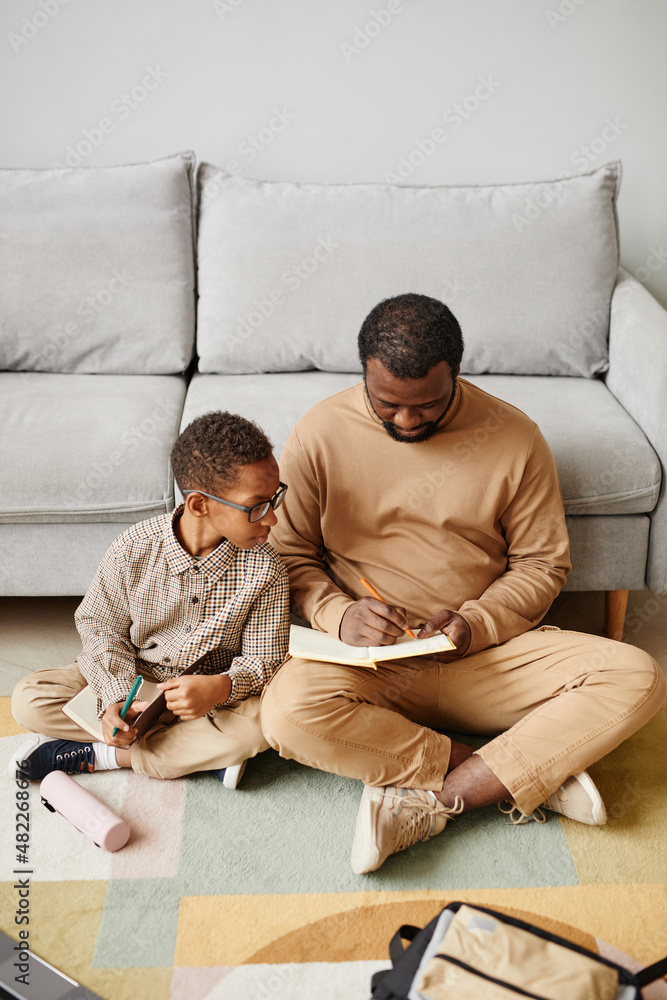 Poster vertical full length portrait of african-american father and son sitting on floor while doing homewo