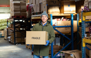 Warehouse worker standing, holding pasteboard box with fragile sign and looking at camera.