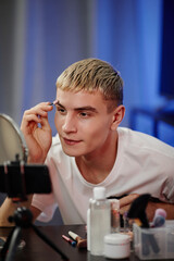 Vertical portrait of blonde young man using makeup while filming male beauty tutorial in studio - obrazy, fototapety, plakaty
