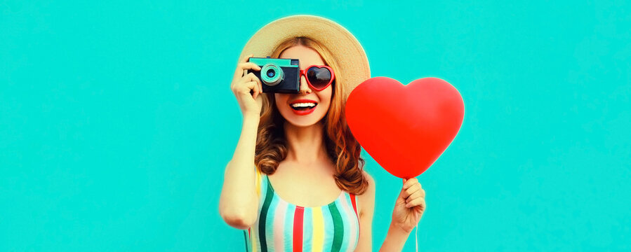 Summer portrait of happy smiling young woman with retro camera and red heart shaped balloon wearing a straw hat on blue background